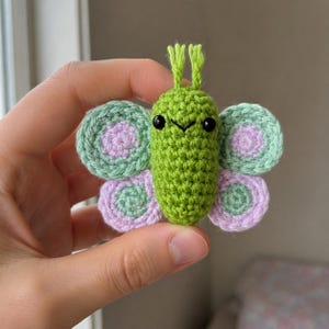 May include: A small crocheted butterfly toy with a bright green body, two antennae, and black bead eyes. The four wings are circular, featuring concentric rings of light green and soft lavender yarn, creating a layered, textured appearance.