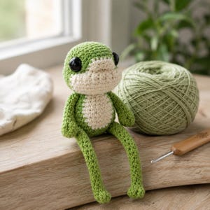Pode incluir: Um pequeno brinquedo de sapo feito à mão em crochê com um corpo verde brilhante, uma barriga cor de creme e grandes olhos redondos de plástico preto. O sapo está posicionado ao lado de um novelo de lã verde claro e uma agulha de madeira.