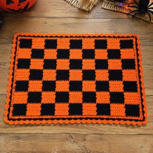 Pode incluir: Um tapete retangular de crochê com um padrão xadrez preto e laranja. O tapete é finalizado com uma borda decorativa festonada laranja que emoldura toda a peça, criando um design de grade consistente em toda a superfície têxtil.