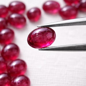 Könnte beinhalten: Ein einzelner ovaler, tiefroter Edelstein, der von einer silberfarbenen Metallpinzette gehalten wird. Der Edelstein hat eine glatte, polierte Cabochon-Oberfläche. Mehrere weitere identische rote ovale Edelsteine liegen unscharf im Hintergrund.