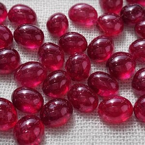Könnte beinhalten: Eine Ansammlung zahlreicher kleiner, glatter, oval geformter Perlen in einer tiefen, leuchtend rubinroten Farbe. Jede Perle hat eine polierte, glänzende Oberfläche, die das Licht reflektiert und helle Glanzlichter auf den runden Steinen erzeugt.