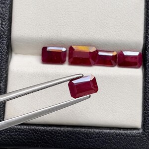 Puede incluir: Una colección de cinco rubíes de color rojo intenso con corte esmeralda. Cuatro de las piedras preciosas están dispuestas en fila sobre una base de exhibición blanca, mientras que un quinto rubí a juego es sostenido por pinzas metálicas plateadas.