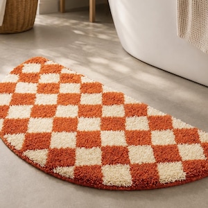Puede incluir: Una alfombra de baño semicircular con un patrón de cuadros repetitivo en color naranja quemado y crema. La alfombra tiene una textura gruesa y copetuda, colocada sobre una superficie plana, mostrando un diseño geométrico distintivo.