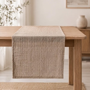 May include: A rectangular table runner featuring a textured, waffle-weave pattern in a natural tan, beige color. The fabric has a visible grid-like structure with finished, stitched edges, draped over the surface of a light-toned wooden table.