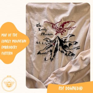 May include: A long-sleeve cream-colored shirt featuring black and red embroidery of a mountain range and a dragon. The text "The Lonely Mountain" is stitched in black thread above the mountain peak. The design includes a small red mark on the mountain.