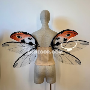 May include: A pair of large, translucent insect-style wings featuring orange, black, and white patterns with distinct vein details, attached to a beige corset worn on a mannequin. The text OSDECOR.COM is visible across the center of the wings.