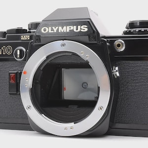Puede incluir: Un cuerpo de cámara de película de 35mm Olympus OM-10 en color negro con montura de lente plateada. El frente muestra la marca Olympus, la designación del modelo OM-10, un indicador rojo de disparo y una superficie de agarre negra texturizada.