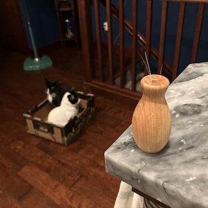 May include: A small, light-colored wooden bud vase with a smooth, rounded body and a narrow neck. The vase features a single small opening at the top, from which two thin, dark, hair-like fibers protrude. The wood grain is visible throughout the surface.