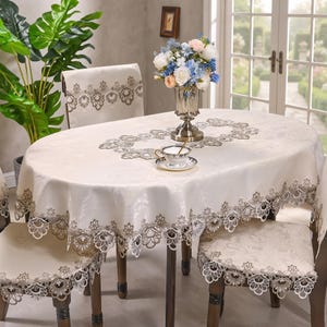 May include: An oval cream-colored tablecloth with an intricate floral lace border and a matching center runner design. The set includes coordinating chair covers with identical lace detailing, a glass vase with a floral arrangement, and a teacup.