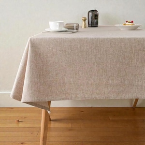 May include: A rectangular table covered with a textured, light beige fabric tablecloth. The table features light-coloured wooden legs. On the surface, there is a white ceramic cup, a small gold-toned metal object, a dark glass bottle, and a white plate.