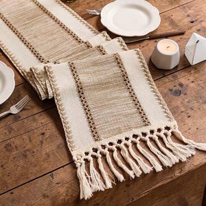 Op de afbeelding: Een rechthoekige tafelloper met een geweven textuur en afwisselende verticale strepen in crème en beige. De tafelloper is aan beide uiteinden afgewerkt met lange, geknoopte crèmekleurige franjes die een decoratieve rand vormen.
