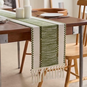 Puede incluir: Un camino de mesa rectangular con un panel central tejido en color verde oliva flanqueado por bordes de tela color crema. El camino incluye detalles decorativos de costura verde a lo largo de los bordes y termina con borlas de flecos color crema.