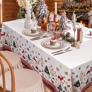 May include: A white rectangular tablecloth featuring a festive pattern of red reindeer, green pine trees, and colourful snowflakes. The fabric has a decorative border along the edges and is set with clear plates, silver cutlery, and cupcakes.