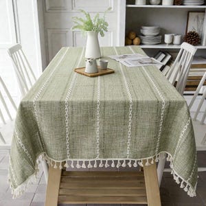May include: A rectangular sage green tablecloth featuring a textured weave, vertical white embroidered stripes, and a decorative fringe trim along the edges. The fabric covers a wooden table, with a white vase and small ceramic items on top.
