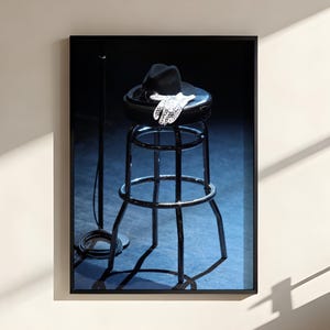 May include: A framed wall art print featuring a black stool with a black fedora hat and white lace gloves resting on the seat. A thin black microphone stand with a coiled cable stands to the left of the stool against a dark blue background.