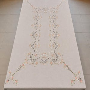 Può includere: Una tovaglia rettangolare color beige caratterizzata da un bordo decorativo centrale con intricati ricami neri simili al pizzo e piccoli accenti floreali rosa. Il disegno si estende verso gli angoli con motivi floreali simmetrici.