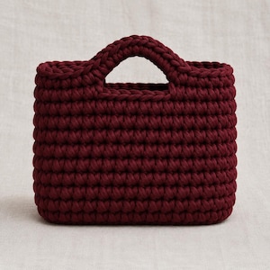 May include: A deep burgundy handbag crafted from thick, chunky yarn in a dense crochet stitch pattern. The bag features a rectangular body with a rounded top edge and two integrated, arched handles formed by the same continuous crochet work.