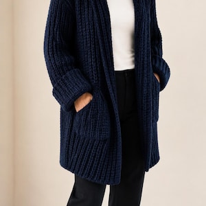 Puede incluir: Un cárdigan con capucha de color azul marino con una textura de punto acanalado grueso, mangas largas con puños anchos y dos grandes bolsillos de parche en la parte delantera. La prenda se lleva sobre una camiseta blanca lisa y pantalones oscuros.