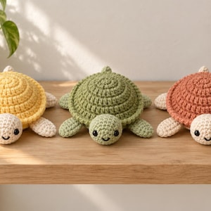 May include: Three handmade crochet turtle plushies arranged in a row. The left turtle is yellow with cream-colored limbs, the center turtle is sage green, and the right turtle is terracotta orange with cream-colored limbs. Each has black eyes.
