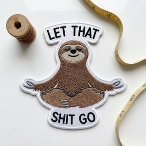 May include: An embroidered patch featuring a brown sloth sitting in a cross-legged position with eyes closed. The text LET THAT is above the sloth and SHIT GO is below it in black letters. A wooden spool and a yellow measuring tape are visible.