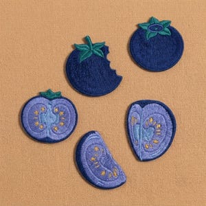 May include: Five embroidered patches shaped like blueberries in various states. One whole berry, one with a bite taken out, and three sliced sections showing light purple interiors with yellow seeds, all featuring dark blue and green thread.