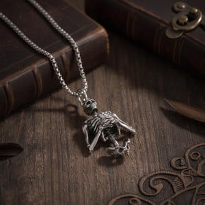May include: A silver-toned metal pendant necklace featuring a detailed skeleton figure. The skeleton is depicted with its arms crossed over its chest, suspended from a thin, textured box chain. The metal has a dark, oxidized finish.