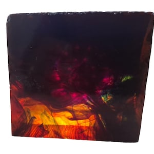 Könnte beinhalten: Ein quadratischer, durchscheinender Harzblock mit einer tiefen, dunklen Farbpalette und wirbelnden internen Mustern in Magenta, Orange und Gelb. Die Oberfläche ist glatt und glänzend, wobei Licht durch die lebendigen, rauchigen und wolkenartigen Pigmente fällt.