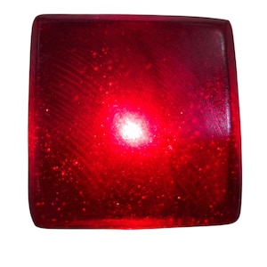 Könnte beinhalten: Ein quadratisches, durchscheinendes rotes Objekt mit einer glatten, leicht abgerundeten Oberfläche. Das Material enthält feine, glitzernde Partikel, und eine helle, konzentrierte weiße Lichtquelle scheint durch die Mitte und erzeugt einen Leuchteffekt.