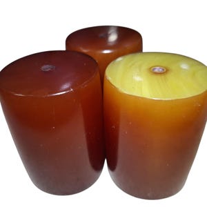 May include: Three cylindrical objects with smooth, polished surfaces. Two of the objects are a deep, translucent reddish-brown colour, while the third object features a lighter, marbled yellow top transitioning into a warm amber-brown base.
