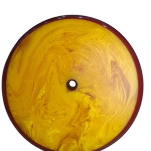 Puede incluir: Un disco circular y plano que presenta un patrón jaspeado de remolinos amarillos vibrantes y marrones dorados. El objeto tiene una superficie lisa y pulida y un borde distintivo de color marrón rojizo oscuro, con un pequeño orificio central.