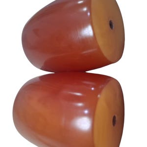 Könnte beinhalten: Zwei glatte, abgerundete, rotbraune Holzobjekte mit glänzender Oberfläche. Jedes Stück hat eine flache, hellere kreisförmige Vorderseite mit einem kleinen, mittigen Loch, was auf funktionale Bauteile wie Möbelknöpfe hindeutet.