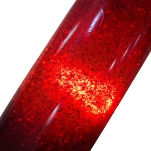 Puede incluir: Un objeto cilíndrico con un acabado rojo profundo y translúcido incrustado con finas partículas de purpurina brillante. La superficie es lisa y brillante, reflejando la luz con un punto de luz concentrado en el centro.