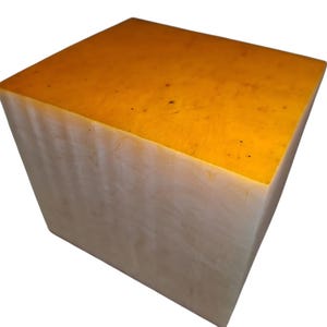Puede incluir: Un cubo sólido con un acabado liso y mate. La superficie superior es de un color naranja ámbar cálido y vibrante con sutiles motas oscuras, mientras que las caras laterales son de un tono blanco crema más claro con un aspecto suave y veteado.