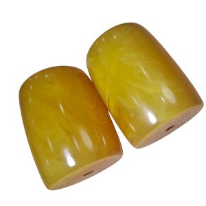 Puede incluir: Dos cuentas cilíndricas con un acabado liso y pulido y un patrón jaspeado en tonos amarillo y ámbar. Cada cuenta presenta un orificio vertical central que atraviesa la longitud de la pieza, mostrando una textura translúcida y arremolinada.