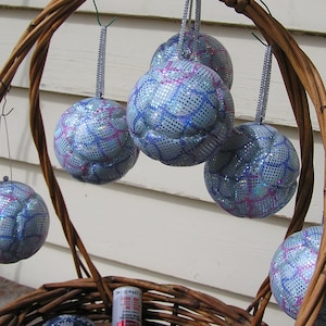May include: Several spherical Christmas baubles with a blue and silver sequin design, suspended by silver ribbons. The decorations are arranged in a woven wicker basket, creating a festive holiday display.