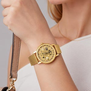 May include: A gold-tone wristwatch featuring a round skeleton dial that reveals the internal mechanical movement. The watch has a fine mesh metal strap and a polished gold-coloured case with Roman numeral hour markers around the outer edge.
