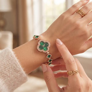 May include: A rose gold-tone watch with a clover-shaped face featuring a green dial and clear stones. The bracelet consists of alternating clover-shaped links with green stones and clear crystals. Two gold-tone rings with filigree patterns.