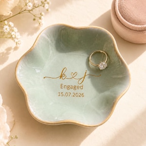 May include: A sage green, flower-shaped ceramic ring dish with gold trim. The center features gold script text reading k & j, the word Engaged, and the date 15.07.2026. A gold engagement ring with a clear stone rests inside the dish.
