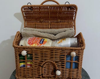 May include: A brown woven wicker basket with a hinged lid, featuring a house-like design with a small arched door and white wooden bead accents. Inside, the basket contains a folded beige towel, a bottle of Softsoap, and other items.