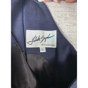 Pode incluir: Uma peça de roupa azul-escura com uma superfície tecida texturizada e um forro castanho-escuro. Uma etiqueta retangular branca está cosida no tecido, com o texto em caligrafia preta Linda Segal, LINDA SEGAL FOR ASHLEY, INC. e tamanho 6.