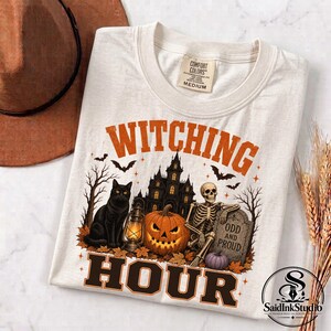 May include: A cream-colored t-shirt featuring a graphic with the text WITCHING HOUR. The design includes a black cat, a lit lantern, a jack-o'-lantern, a skeleton leaning on a tombstone that reads ODD AND PROUD, a haunted house, and bats.