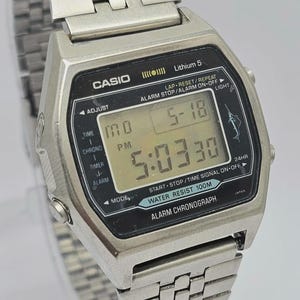 May include: A silver-tone Casio digital watch with a metal link band. The rectangular face features a black display with white text including CASIO, Lithium 5, and WATER RESIST 100M. The digital screen shows the time 5:03:30, date 5-18, and a marlin icon.