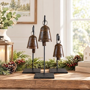 May include: Three rustic metal bells with a weathered brown finish, each suspended from a black metal hook on a thin vertical rod attached to a square black base. The bells vary in height, with the center bell being the tallest of the set.