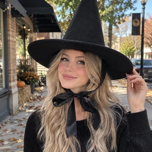 May include: A black pointed hat with a wide brim and a black satin ribbon tie that fastens under the chin. The hat features a tall, conical crown and a smooth, dark fabric texture, designed to be worn on the head with the ribbon tied in a bow.