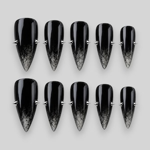 Può includere: Un set di dieci unghie finte a stiletto con una finitura nera lucida che sfuma in un motivo argentato crepato sulle punte. Ogni unghia è decorata con due piccole borchie metalliche argentate e appuntite sui lati.