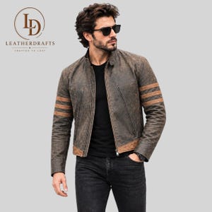 Può includere: Una giacca in pelle marrone effetto invecchiato con tre strisce orizzontali color cuoio su ogni manica, colletto alla coreana, chiusura frontale con zip e un pannello in pelle color cuoio sul fondo. Indossata sopra una t-shirt nera e jeans neri.