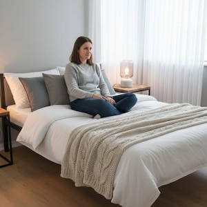May include: A bed featuring a white duvet cover, a thick beige cable-knit throw blanket, and several pillows in white and grey. The bed has a black metal frame and is positioned in a room with light wood flooring and white curtains.