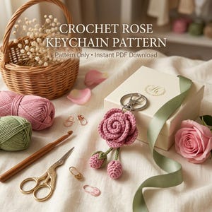 May include: A crochet rose keychain pattern with text reading CROCHET ROSE KEYCHAIN PATTERN Pattern Only Instant PDF Download. Includes a pink crocheted rose keychain, a wicker basket, pink and green yarn, a wooden crochet hook, and scissors.