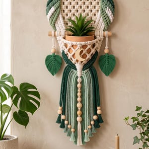 May include: A macrame wall hanging shaped like an owl, crafted from cream, light green, and dark green cord. It features large black and white eyes, a central terracotta pot holder with a small succulent, and a gold crescent moon charm.