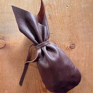 May include: A dark brown leather pouch with a rounded base and a gathered top. The opening is secured by a thin leather cord wrapped around the neck, creating two pointed, ear-like flaps extending upward from the cinched closure.